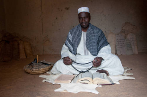 Dark-skinned man in white robe sits before two manuscripts, Qur’an boards lean against wall behind