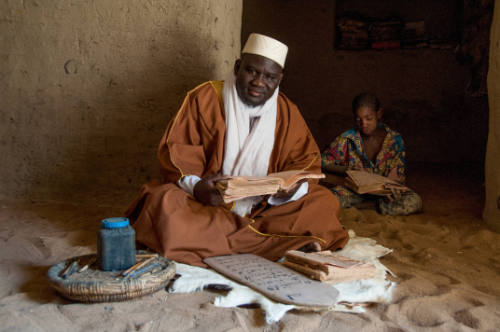 Dark-skinned seated man looks up from a manuscript while a seated child reads a manuscript behind