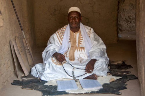 Dark-skinned man in embroidered white robe holds prayer beads and sits before an open manuscript