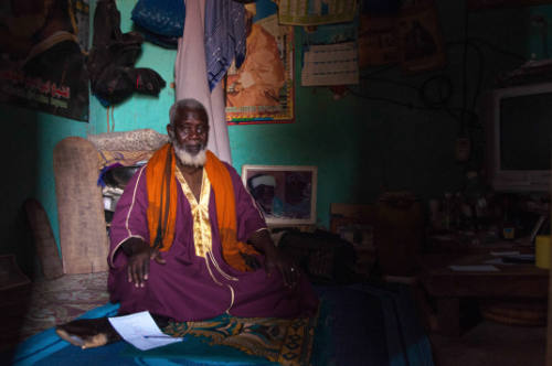 Dark-skinned man with white hair, beard, purple robe sits on mat in dim room, green wall