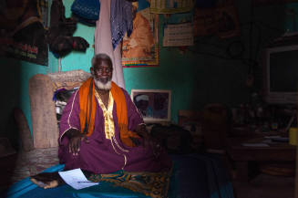 Dark-skinned man with white hair, beard, purple robe sits on mat in dim room, green wall