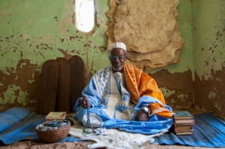 Dark-skinned man in cap, glasses, blue robe, orange shawl sits beside stacked books,Qur’an boards