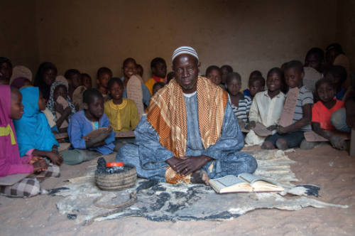 Dark-skinned man sits cross-legged with manuscript as boys and girls with Qur’an boards sit behind