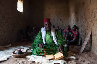 Dark-skinned man in green robe sits in dim room with prayer beads & manuscript with students behind