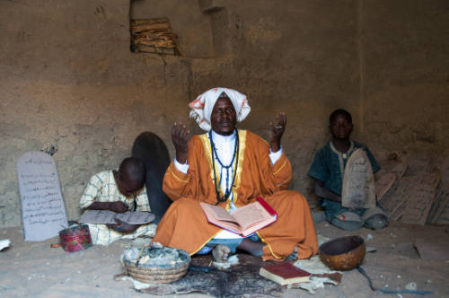 Dark-skinned, robed man with hands raised & manuscript on lap sits between boys with Qur’an boards