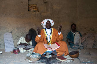 Dark-skinned, robed man with hands raised & manuscript on lap sits between boys with Qur’an boards