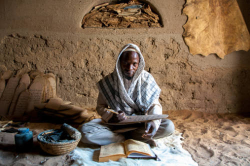 Dark-skinned man sits copying from manuscript onto Qur’an board with additional Qur’an boards behind