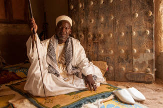 Dark-skinned man in white robe and cap sits on a mat before a large wooden door with metal fittings