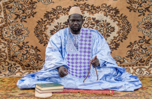 Dark-skinned man in embroidered robe sits on mat with manuscripts in front of hanging floral carpet