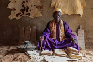 Dark-skinned man in purple robe sits cross-legged on animal hide with manuscripts and Qur’an boards