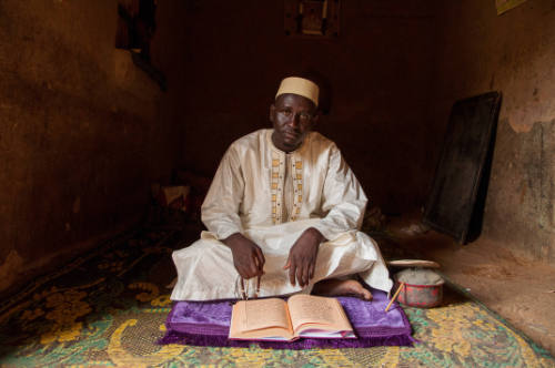 Dark-skinned man in white robe sits cross-legged before a manuscript on a mat in a dark interior