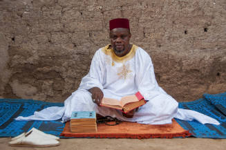 Dark-skinned man in cap and white robe sits on mat holding a manuscript while another sits in front