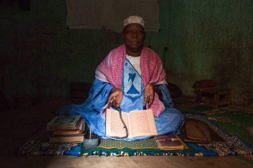 Dark-skinned man in robe sits on mat in dark interior with manuscript on lap & other books nearby 