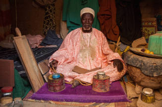 Dark-skinned man in coral robe sits on cushion in crowded interior with manuscript on lap