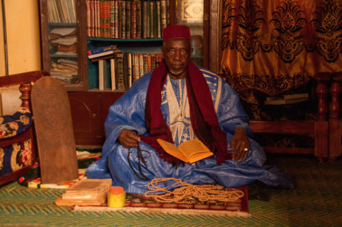 Dark-skinned man in cap & indigo robe sits with manuscript on lap & Qur’an board to right