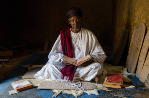 Dark-skinned man in white robe sits cross-legged in dark interior with manuscript and Qur’an boards