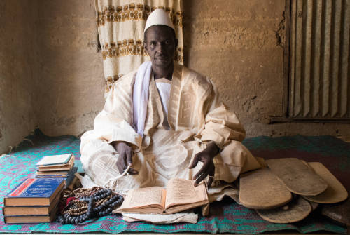 Dark-skinned man in white robe and cap sits cross-legged with a manuscript, beads, and Qur’an boards