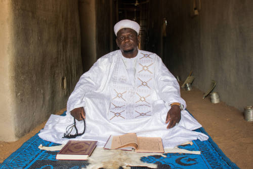 Dark-skinned man in white robe & cap sits cross-legged on blue mat in dim interior with manuscripts