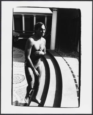 Black-and-white full-body photo of man in swim briefs walking up steps with hand covering chest