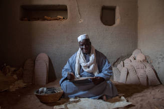 Dark-skinned man in robe sits on animal hide writing on Qur’an board with other boards behind