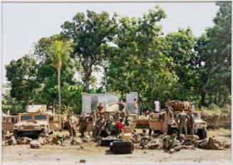 People lounge around 3 army vehicles in a sandy area with trees in the background.