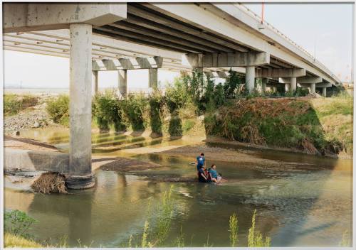 Group of people stand and sit in the middle of a shallow river under a highway bridge.