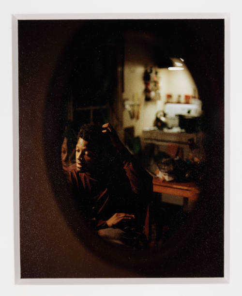 Oval mirror shows Black male-presenting figure seated hand to head at vanity in dimly lit room.