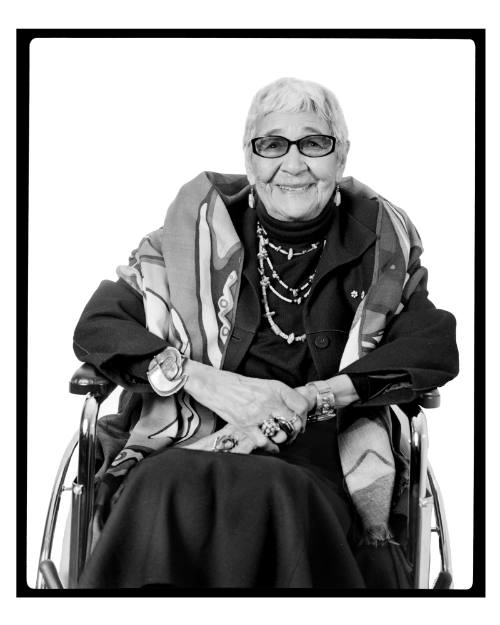 Portrait of woman seated in a wheelchair wearing a patterned shawl, multi-strand necklace and rings