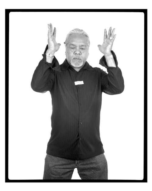 Black-and-white photo of man with medium skin tone, standing with raised hands and a downward gaze