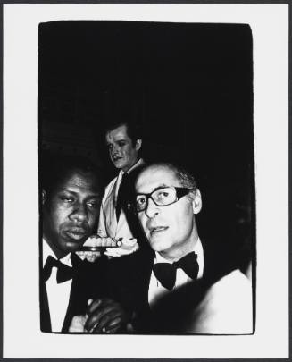 Two men wearing formal wear, one wearing thick black framed glasses; a waiter is in the background