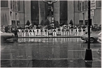 A crowd of people in the rain hold posters spelling the phrase “LEGALIZE ABORTION”.