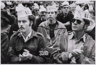 Two stern male-presenting people in white paper caps reading "G.I." clap among a crowd of others.