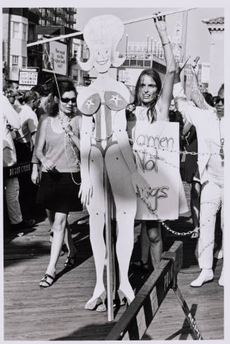 2 female-presenting people lead a crowd with a cut out of a scantily clad woman in chains.