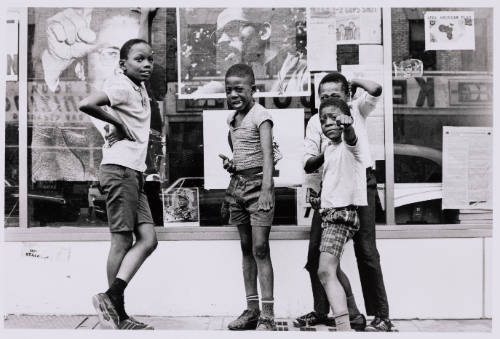 4 Black children stare into the camera while in front of a store window covered in photos.