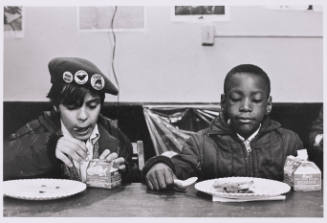 Two children, one Latine and one Black, eat breakfast side by side off of paper plates.