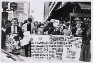 Three people lead a crowd holding a poster with small portraits of dead soldiers.