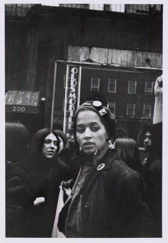 A female-presenting person wearing a beret with pins looks past the camera in the midst of a crowd.