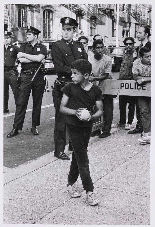 A child palming a baseball walks before a police line with a crowd and several police officers.