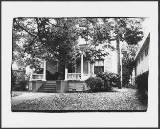 Tree covering a view of a late 19th or early 20th century clapboard-sided house with porch 