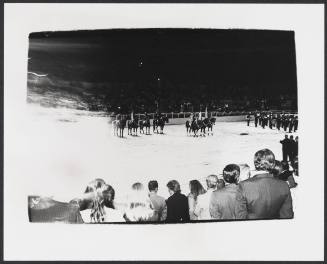 People standing in bleachers overlooking an arena with a marching band and men on horseback