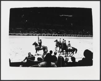 Audience view of an indoor arena with a rider carrying a flag on horseback followed by four riders 