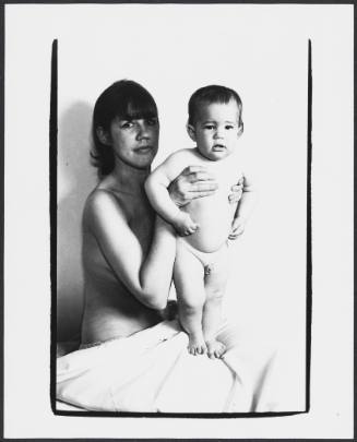 Woman with light skin tone and cloth draped over her lap steadies a standing naked baby on her lap