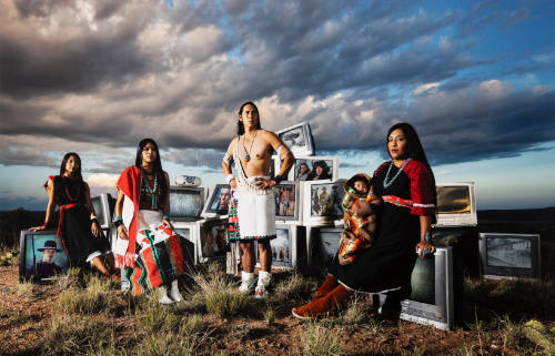 Three women, a man and a baby in Pueblo attire pose amidst discarded televisions in arid landscape