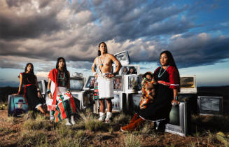 Three women, a man and a baby in Pueblo attire pose amidst discarded televisions in arid landscape