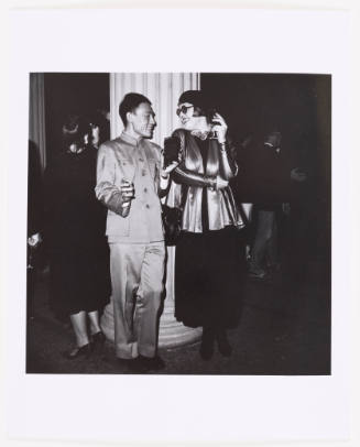 Black-and-white photo of man in Mao suit talking to woman wearing silk jacket, beret, and sunglasses