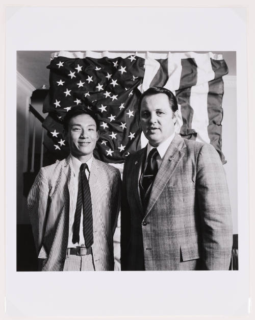 Black-and-white photo of two men wearing suits and ties posing in front of a wrinkled American flag