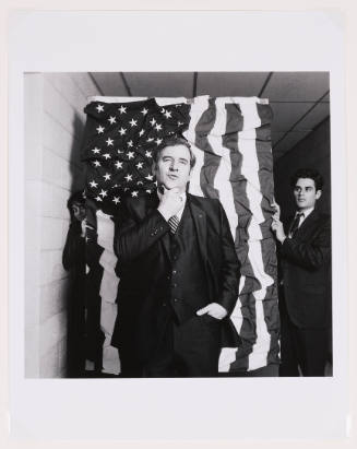 Black-and-white photo of a light-skinned man in suit in front of wrinkled American flag 