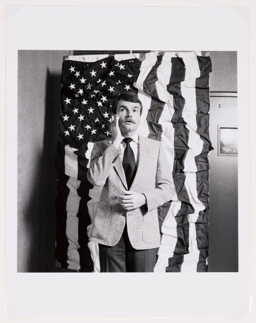 Black-and-white photo of a man with mustache wearing a suit in front of a wrinkled American flag