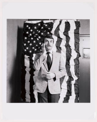 Black-and-white photo of a man with mustache wearing a suit in front of a wrinkled American flag