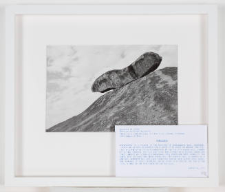 Black-and-white photo of a large, oblong boulder perched on a cliff with clouds in background